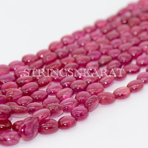 May include: A strand of oval-shaped, pink ruby beads. The beads are smooth and polished, with a slight sheen. The beads are strung on a clear thread. The text "STRINGSNKARAT" is visible in the image.
