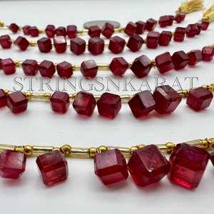May include: A strand of red gemstone beads with a gold spacer between each bead. The beads are faceted and have a square shape. The strand is on a white surface.