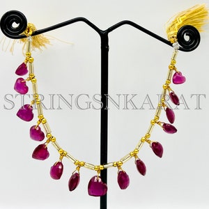 May include: A gold and silver necklace with a row of dangling heart-shaped purple gemstones. The necklace is displayed on a black stand with a white background. The text "STRINGSNKARAT" is visible in the image.