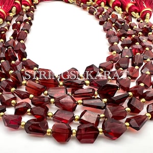 Puede incluir: Una hebra de cuentas de piedras preciosas de color rojo intenso con forma irregular facetada. Las cuentas están enhebradas en un alambre de color dorado y tienen un borla en cada extremo.