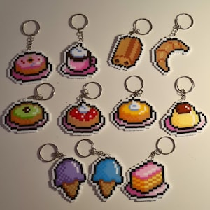 Op de afbeelding: Een verzameling pixel art sleutelhangers met verschillende voedselitems. De sleutelhangers bevatten een donut, koffiekopje, croissant, brood, cake, ijsje en pudding. Elke sleutelhanger is gemaakt van kleurrijke kralen en bevestigd aan een zilveren sleutelhanger.