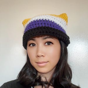 May include: A handmade crochet hat with cat ears, featuring the non-binary pride flag colors: yellow, white, purple, and black. The hat is worn by a person with dark hair and a neutral expression. The hat is a unique accessory.