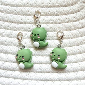 May include: Three green dinosaur-shaped charms with silver-colored clasps. Each charm has a white belly and pink cheeks. The charms are arranged on a white, textured surface.