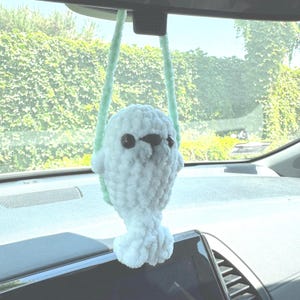 May include: A handmade, white crocheted seal car charm with black eyes and a light green hanging cord. The plush seal is hanging from a car's rearview mirror, adding a touch of whimsy to the vehicle's interior.
