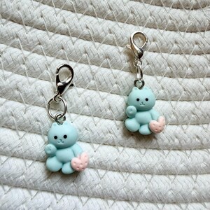 May include: Two light blue cat charms with silver-colored clasps. Each charm features a small, light blue cat holding a pink heart. The charms are designed for use as accessories, such as zipper pulls or keychain attachments.