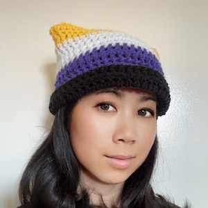 May include: A handmade crochet hat featuring the non-binary pride flag colors: yellow, white, purple, and black. The hat has a brim and a pointed top. The hat is worn by a person with dark hair.