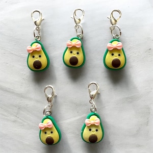 Könnte beinhalten: Fünf avocadoförmige Anhänger mit silberfarbenen Verschlüssen. Jeder Anhänger hat eine grüne Schale, gelbes Fruchtfleisch und einen braunen Kern. Eine rosa Schleife ist oben an jeder Avocado befestigt. Diese Anhänger eignen sich für die Schmuckherstellung oder als dekorative Accessoires.