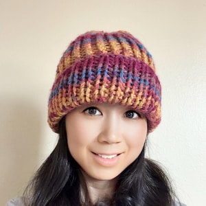 May include: A chunky knit beanie featuring a variegated yarn pattern in shades of magenta, mustard yellow, and blue. The hat is constructed with a thick ribbed texture and includes a wide, folded cuff that sits securely around the crown of the head.