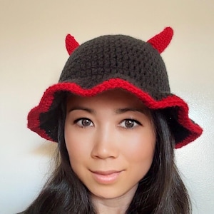 May include: A handmade crochet hat in dark gray with a red ruffled brim and two red horns. The hat is a unique accessory, perfect for costumes or adding a playful touch to any outfit. The hat is a fun and quirky fashion statement.