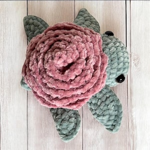 May include: A handmade plush turtle with a large, rose-shaped shell. The shell is a soft, dusty rose color, while the turtle's body and flippers are a muted teal. The turtle has black button eyes and is made of a textured, plush material.