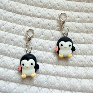 May include: Two small penguin charms with silver-colored clasps. The penguins have black bodies, white bellies, and yellow feet. They have pink accents on their wings and are attached to a silver-colored clasp.