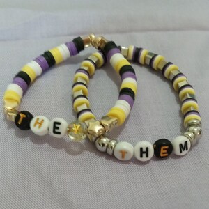 May include: Three bracelets with yellow, black, and purple beads. The bracelets spell out "THE" and "THEM" in white letters.