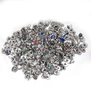 Puede incluir: Un gran mont&oacute;n de anillos plateados con varios engastes de piedras preciosas. Los anillos presentan una variedad de piedras de colores, incluyendo azules, rojas, verdes y rosas, en diferentes formas y tama&ntilde;os. Los anillos se muestran sobre un fondo blanco.