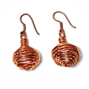 May include: A pair of copper wire earrings. Each earring has a spiral design with a small, round, wire-wrapped orb at the centre. The earrings have a hook closure and are shown against a white background.