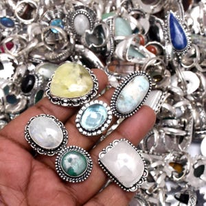 Puede incluir: Una colecci&oacute;n de anillos de plata con diversos engastes de piedras preciosas. Los anillos presentan piedras en tonos de azul, verde, amarillo y blanco. Los anillos se muestran en una mano, con muchos m&aacute;s anillos visibles en el fondo.