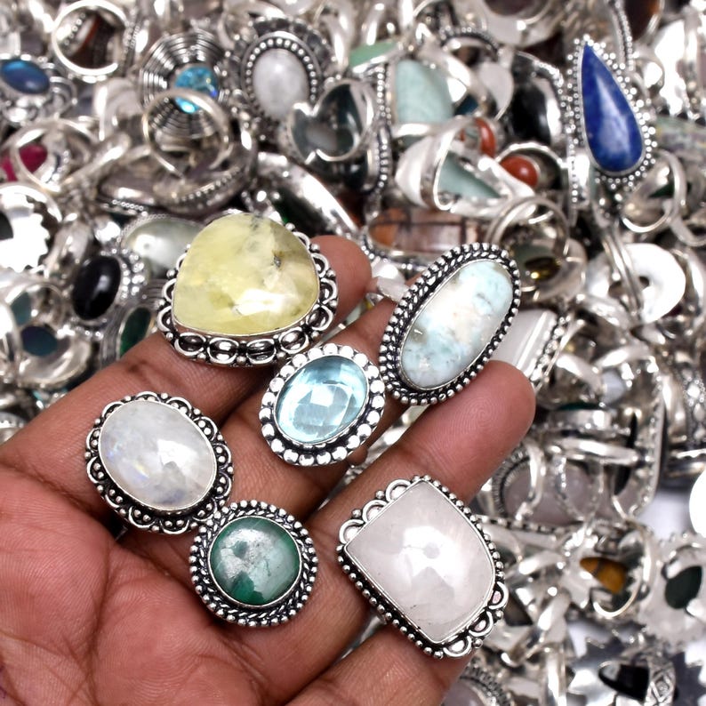 Puede incluir: Una colecci&oacute;n de anillos de plata con diversos engastes de piedras preciosas. Los anillos presentan piedras en tonos de azul, verde, amarillo y blanco. Los anillos se muestran en una mano, con muchos m&aacute;s anillos visibles en el fondo.