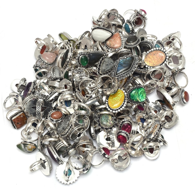 Puede incluir: Un gran mont&oacute;n de anillos de plata con diversos engastes de piedras preciosas. Los anillos presentan una variedad de piedras de colores, incluyendo rojo, verde, amarillo y azul, en diferentes formas y tama&ntilde;os. Los anillos se muestran sobre un fondo blanco.