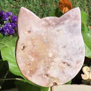 May include: A light pink and tan cat-shaped crystal carving. The stone has a natural, textured appearance with small cavities. The carving is mounted on a thin, gold-colored metal stand, with a blurred background of greenery and flowers.