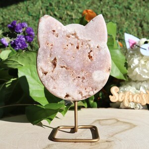 May include: A pink cat-shaped crystal on a gold metal stand. The crystal has a rough, textured surface.