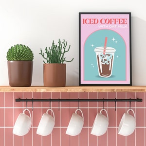 Op de afbeelding: Een zwart ingelijste poster met een roze achtergrond en een blauwe en witte illustratie van een glas met ijskoffie en een rietje. De tekst op de poster luidt "ICED COFFEE WITH OAT MILK".