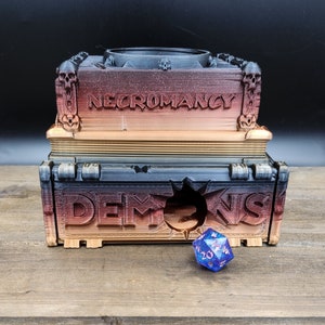 May include: A three-tiered, dark brown, black, and gold painted wooden box with the word "NECROMANCY" printed on the top tier. The box has a hole in the front and a blue and silver dice is sitting in front of the box.