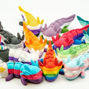 May include: A collection of 14 axolotl figurines, each painted in different colors and patterns representing various pride flags. The axolotls are arranged in a circle on a white background.