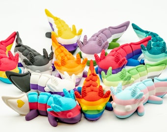 May include: A collection of 14 axolotl figurines, each painted in different colors and patterns representing various pride flags. The axolotls are arranged in a circle on a white background.