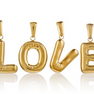 Może przedstawiać: Złoty naszyjnik z zawieszką z czterema literowymi zawieszkami, które tworzą słowo "LOVE". Każda litera jest oddzielnym zawieszką i ma lekko napompowany, balonowy wygląd.
