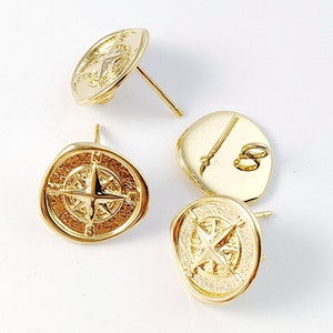 Puede incluir: Cuatro pendientes de brújula de color dorado con un diseño de estrella. Los pendientes están sobre un fondo blanco.