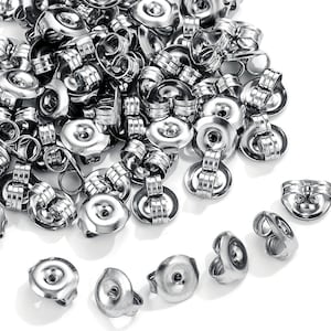 May include: A pile of shiny, silver-colored earring backs, also known as butterfly backs or friction backs, are scattered on a white surface. The earring backs are made of metal and have a post-and-clasp design for securing earrings.