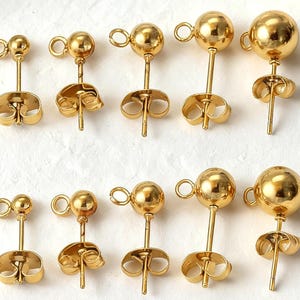 May include: A collection of ten gold-toned metal stud earrings arranged in two rows. Each earring features a spherical ball bead attached to a small circular loop and includes a matching butterfly-style earring back for secure fastening.
