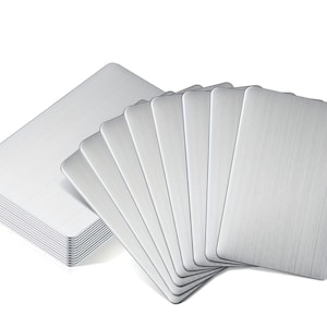 Puede incluir: Un conjunto de tarjetas metálicas plateadas con esquinas redondeadas. Una pila de tarjetas junto a un grupo similar en abanico. Las tarjetas tienen una textura de metal cepillado, probablemente para juegos o elementos decorativos.