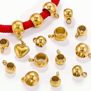 May include: A collection of gold-coloured jewellery findings, including round beads, open-ended cups, and a heart-shaped charm. The findings are displayed on a white surface and a red cord. The beads and cups have small loops for attaching to other jewellery components.