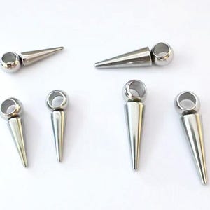 May include: Six silver-toned metal charms, each featuring a smooth, conical spike attached to a circular ring. The spikes vary slightly in length and thickness, with a polished, reflective finish. These components are arranged in a loose group.