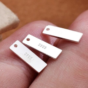 Puede incluir: Primer plano de tres pequeños dijes rectangulares plateados con un agujero en un extremo. Los dijes tienen el texto "S925" estampado. Los dijes se muestran sobre un fondo rosado.
