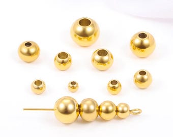 May include: A collection of shiny, gold-colored metal beads in various sizes. Some beads have a hole through the center, and a few are strung together on a metal pin. The beads are arranged on a white surface.
