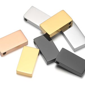 Op de afbeelding: Een verzameling rechthoekige metalen hangers in verschillende kleuren, waaronder zilver, goud, roségoud en zwart. Elke hanger heeft een glad, gepolijst oppervlak en een klein gaatje om aan een ketting of koord te bevestigen. De hangers liggen op een witte achtergrond.
