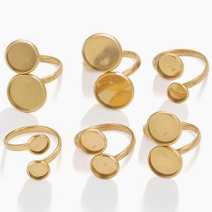 Op de afbeelding: Zes goudkleurige metalen ringen, elk voorzien van twee ronde zettingen voor cabochons of stenen. De ringen hebben een open, verstelbaar bandontwerp met een gladde, matte afwerking. Elk stuk is gerangschikt om de dubbele cupstructuur te tonen.