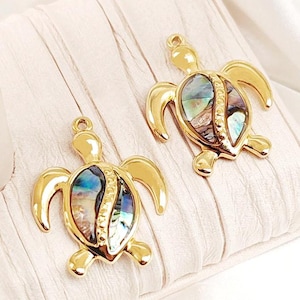 May include: Two gold-tone turtle charms with iridescent abalone shell inlays. The charms are detailed with a textured shell design and have a loop for attaching to jewelry.