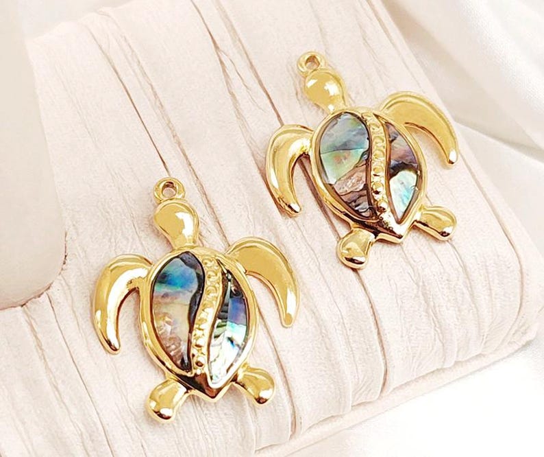 May include: Two gold-tone turtle charms with iridescent abalone shell inlays. The charms are detailed with a textured shell design and have a loop for attaching to jewelry.