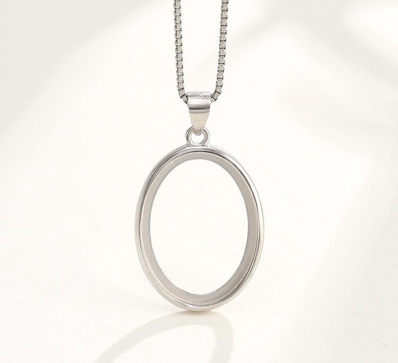 Puede incluir: Un collar con un colgante ovalado de plata y una cadena. El colgante es de forma ovalada simple con una superficie lisa.