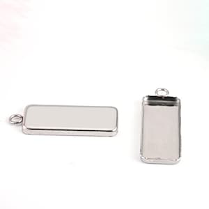 Puede incluir: Dos bandejas de colgante de metal plateadas con forma rectangular y un pequeño bucle en la parte superior para sujetar a una cadena o cordón. Las bandejas están diseñadas para contener objetos pequeños, como resina, vidrio u otros materiales.