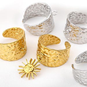 Peut inclure: Gros plan de plusieurs bagues réglables en or et argent. Les bagues ont une texture martelée et une petite boucle pour attacher des breloques. Une bague dorée a un charm en forme de soleil.