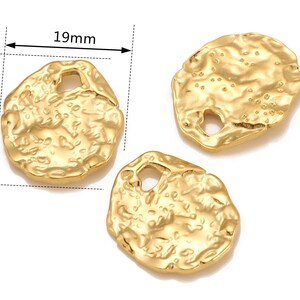 Może przedstawiać: Dwa złote metalowe charms o nieregularnej, fakturowanej powierzchni. Każdy charm ma otw&oacute;r w środku. Charms mają średnicę około 19 mm.