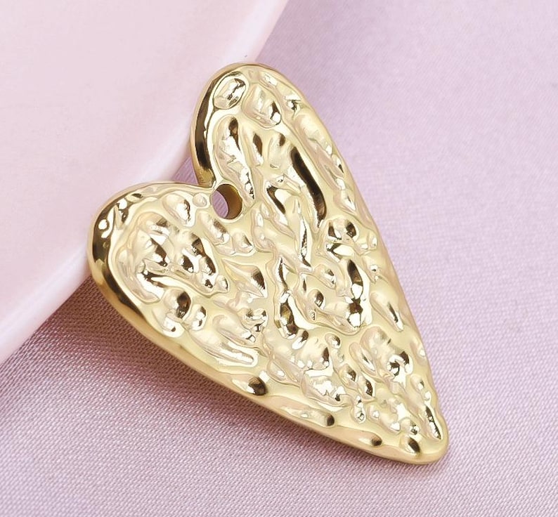 May include: A gold heart-shaped pendant with a textured surface. The pendant has a detailed design with a raised pattern.