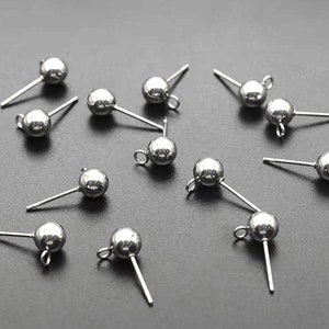 Op de afbeelding: Een close-up van zilveren oorbelpennen met ronde ballen aan het uiteinde. De oorbelpennen zijn gerangschikt op een grijs oppervlak.