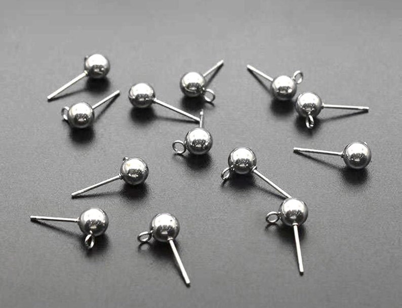 Op de afbeelding: Een close-up van zilveren oorbelpennen met ronde ballen aan het uiteinde. De oorbelpennen zijn gerangschikt op een grijs oppervlak.