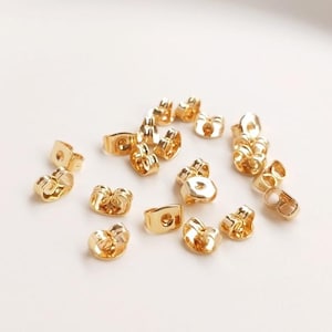 Puede incluir: Primer plano de un grupo de respaldos de pendientes de color dorado. Los respaldos están hechos de metal y tienen un pequeño agujero en el centro. Están diseñados para ser usados con pendientes de poste.