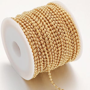 Op de afbeelding: Een spoel met goudkleurige kogelketting. De ketting is strak gewikkeld rond een witte plastic spoel. De ketting is gemaakt van kleine, gelijkmatig verdeelde, ronde kralen. De ketting heeft een heldere, glanzende goudkleur.