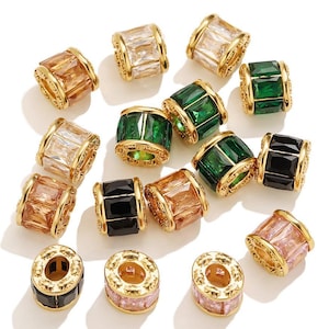 Op de afbeelding: Een verzameling goudkleurige metalen kralen met rechthoekige en vierkante edelstenen in verschillende kleuren. Kleuren zijn helder, groen, zwart, roze en amber. De kralen hebben een cilindrische vorm.